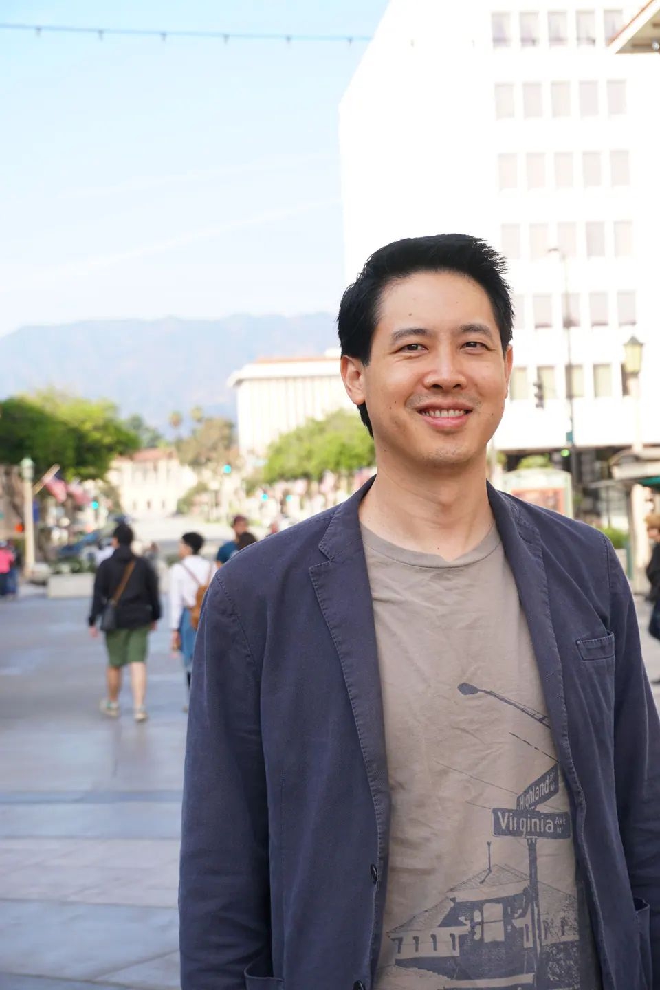 A smiling man in a blue blazer stands on a busy pedestrian plaza, with faint mountains in the distance behind him.