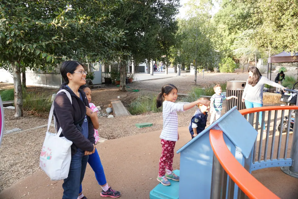 In an outdoor play garden under trees, a woman with a tote bag watches as a girl in a striped shirt points at a water-play exhibit.