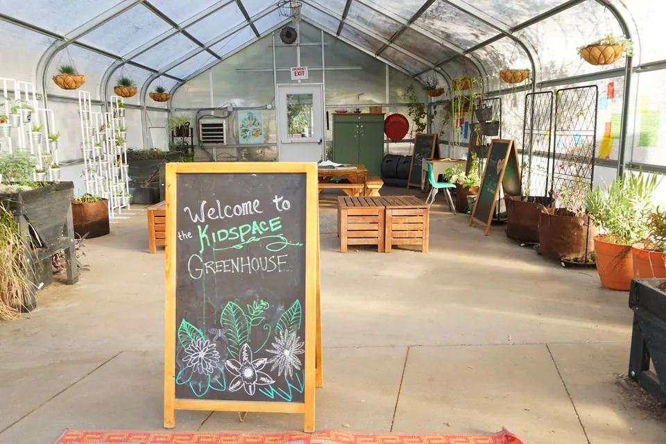 A chalkboard sign reading 'Welcome to the Kidspace Greenhouse' inside a plastic-roofed greenhouse with potted plants and wooden benches.