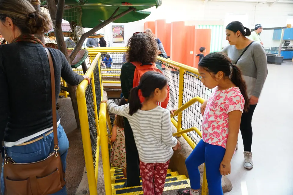 Two girls climb the yellow-railed stairs of an indoor climbing structure among other families.
