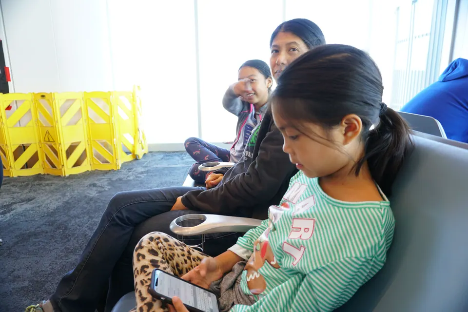 At an airport gate, a girl in a green striped top plays on a phone next to a woman, with another girl in the seat beyond and yellow barriers by the window.