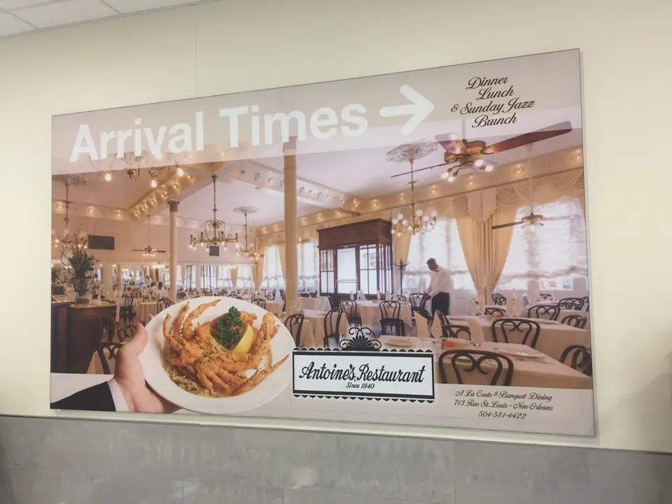 An 'Arrival Times' advertisement for Antoine's Restaurant on an airport wall, showing a chandeliered dining room and a plate of seafood.