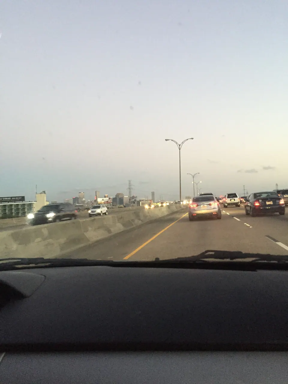 The view through a car windshield on a highway at dusk, with a city skyline low on the horizon.