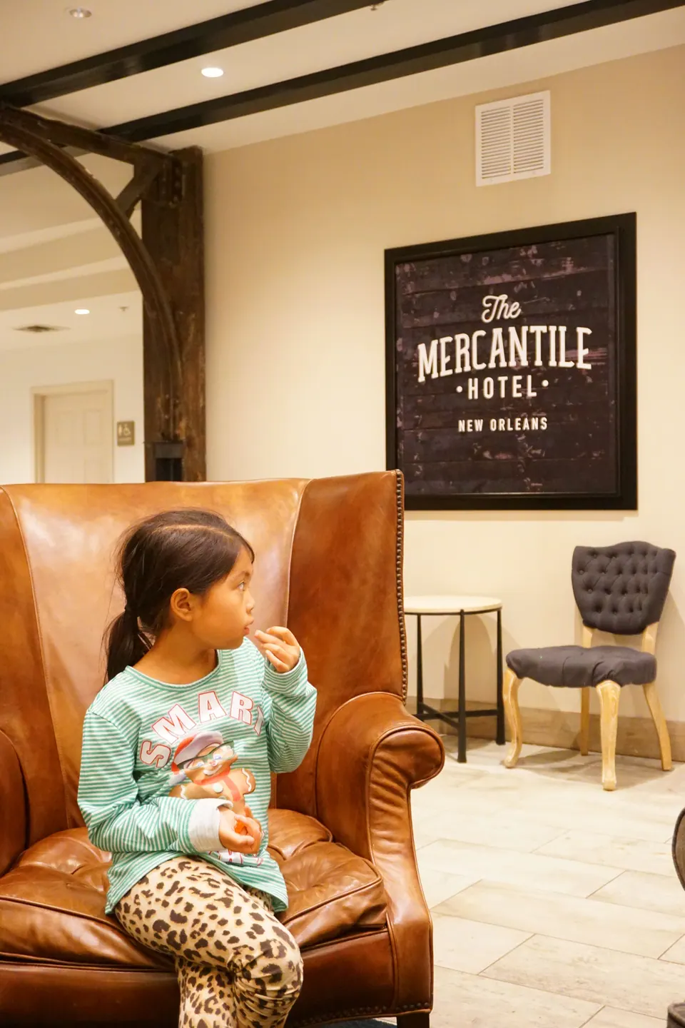 A girl in a green striped top and leopard leggings sits in a brown leather wing chair beneath a framed sign reading The Mercantile Hotel New Orleans.