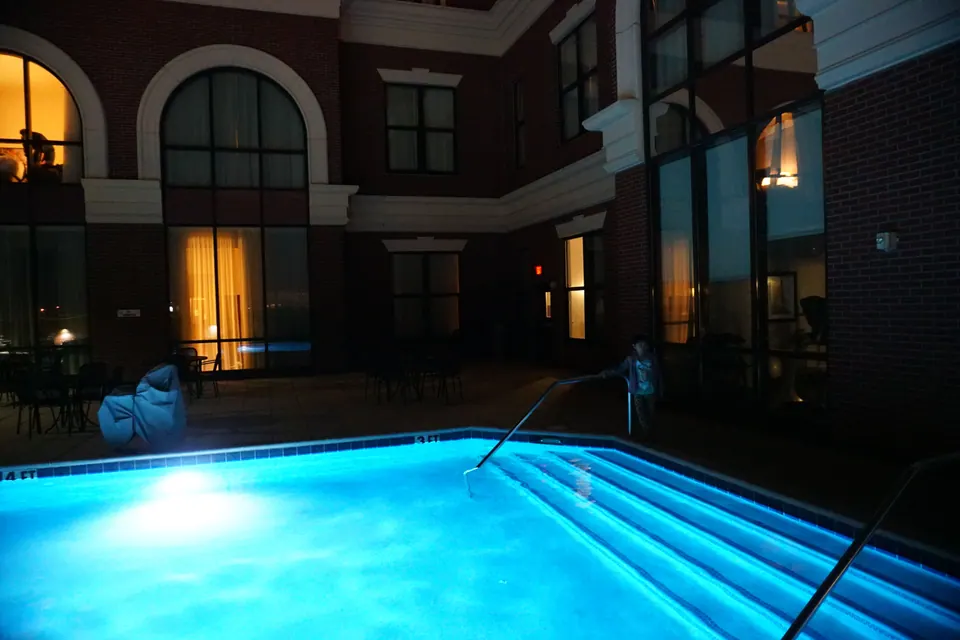A glowing blue rooftop pool at night beside brick hotel walls with arched windows, a child standing by the steps.