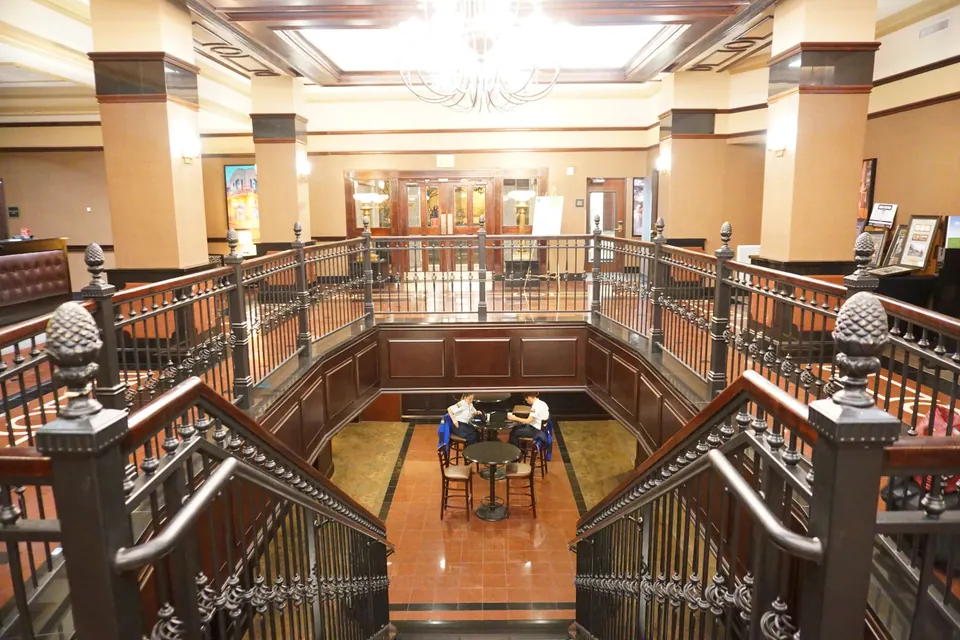 Looking down an ornate double staircase with iron railings into a hotel lobby with tall columns and a chandelier.