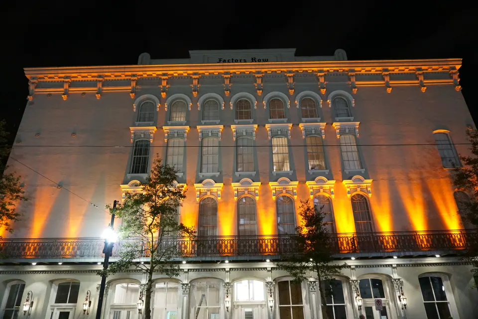 The tall facade of the Factors Row building at night, washed in orange uplighting, with an iron balcony along its lower floor.