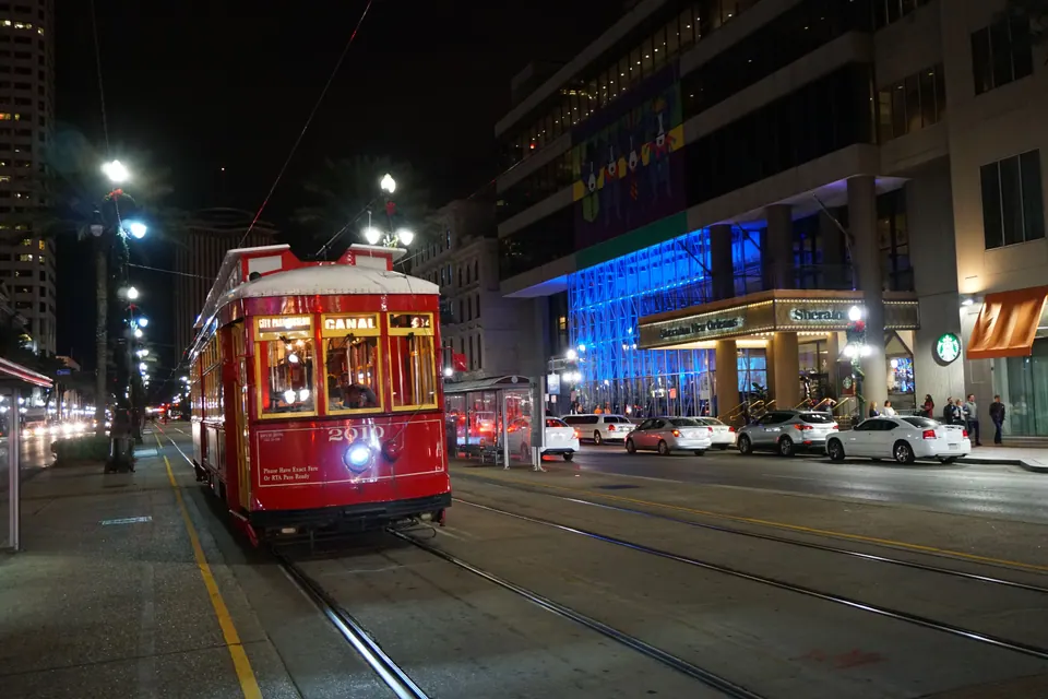 A red streetcar with 'Canal' on its sign glows at its stop at night, with hotel lights and parked cars behind.