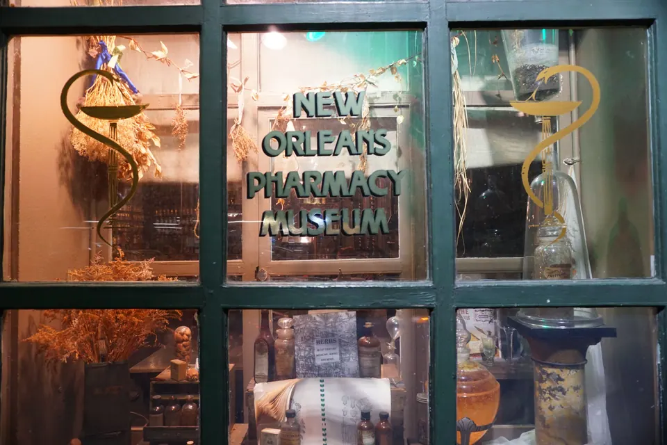 Gold lettering reading New Orleans Pharmacy Museum on a paned window, with old apothecary jars and dried herbs behind the glass.