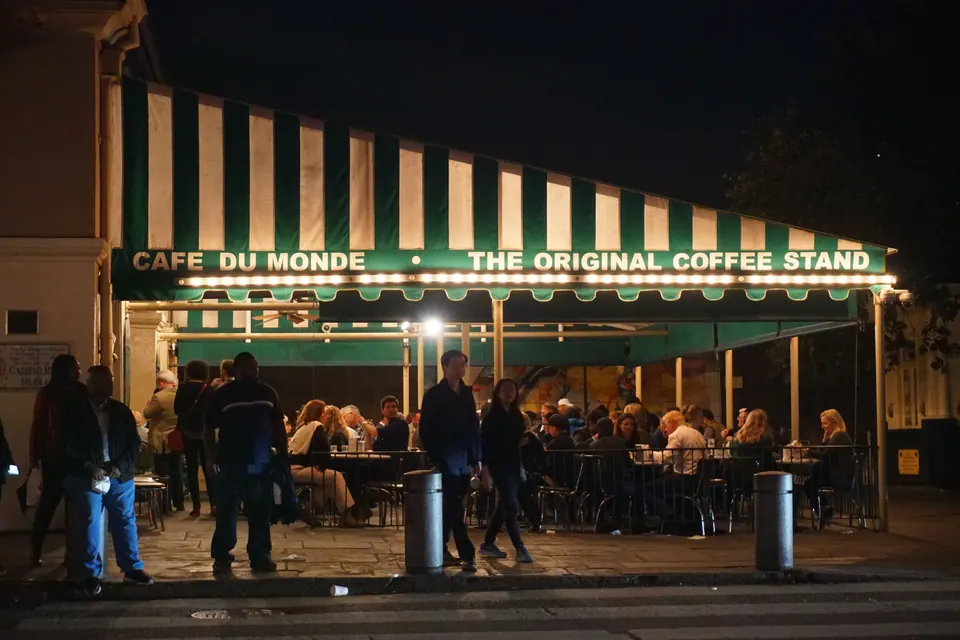 Café du Monde's green-and-white striped awning lit up at night, with the open-air tables full of people.