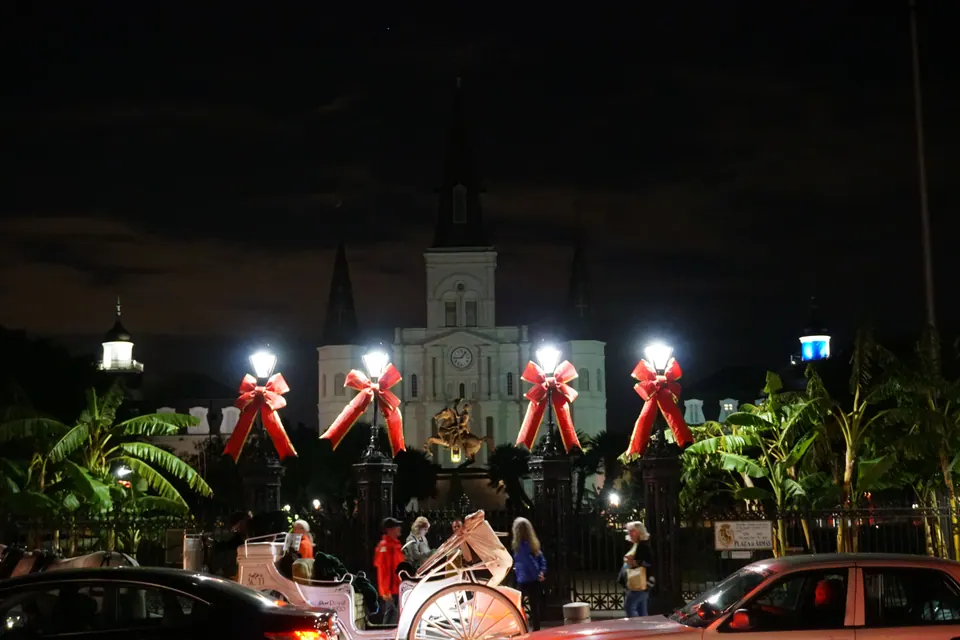 A white cathedral lit at night behind iron gates hung with big red bows, palms on either side and a white carriage passing in front.