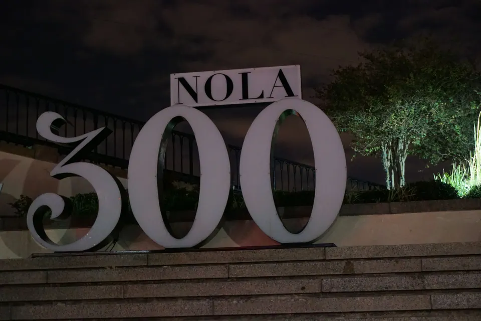 Giant white 'NOLA 300' letters lit at night on stone steps, with an iron railing and trees behind.