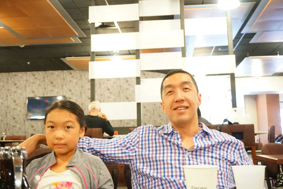 A man in a checked shirt sits with his arm around a girl at a table in a hotel breakfast room, paper cups in front of them.