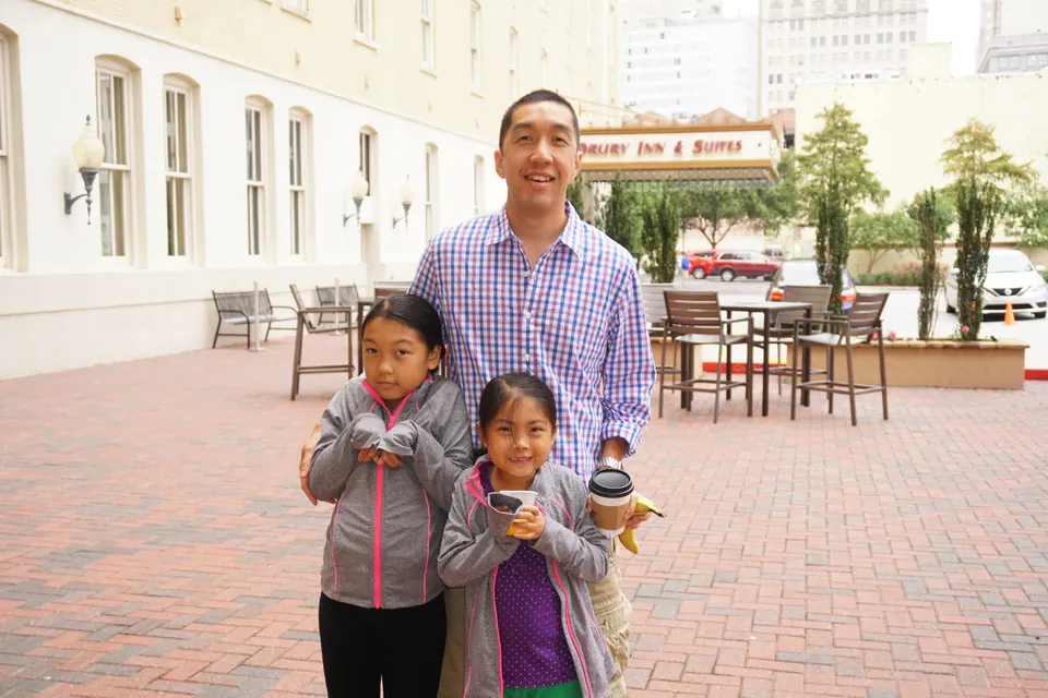 A man in a checked shirt stands with two girls holding snacks and a cup on a brick courtyard, a Drury Inn & Suites sign behind them.