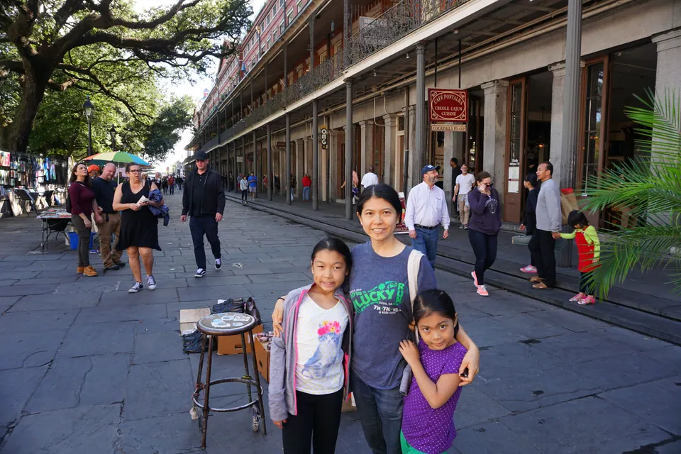 A woman and two girls pose on flagstones under a long arcade with iron galleries above, street vendors and passers-by around them.