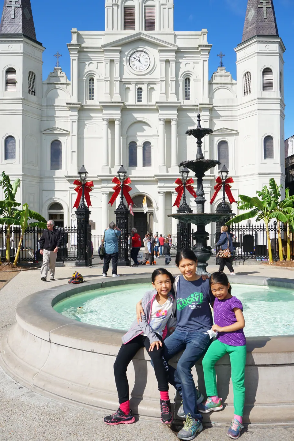 A woman and two girls sit on the rim of a fountain in front of the white, three-spired St. Louis Cathedral under a bright blue sky, red bows on the gates.