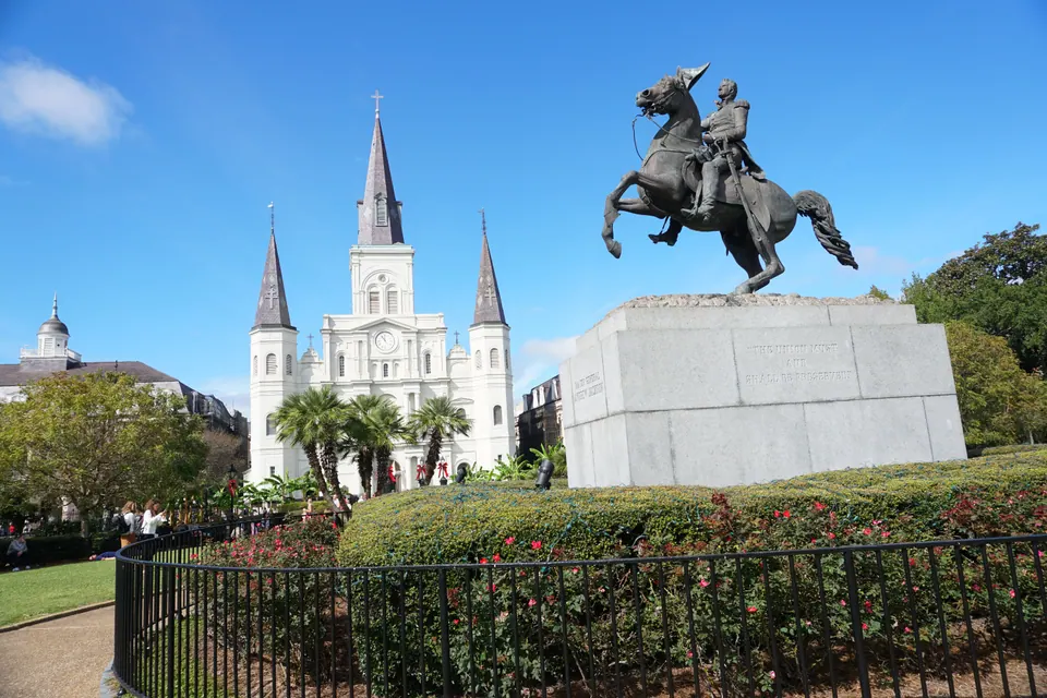 A bronze equestrian statue on a granite base, with the white three-spired cathedral behind a garden and palm trees under a blue sky.