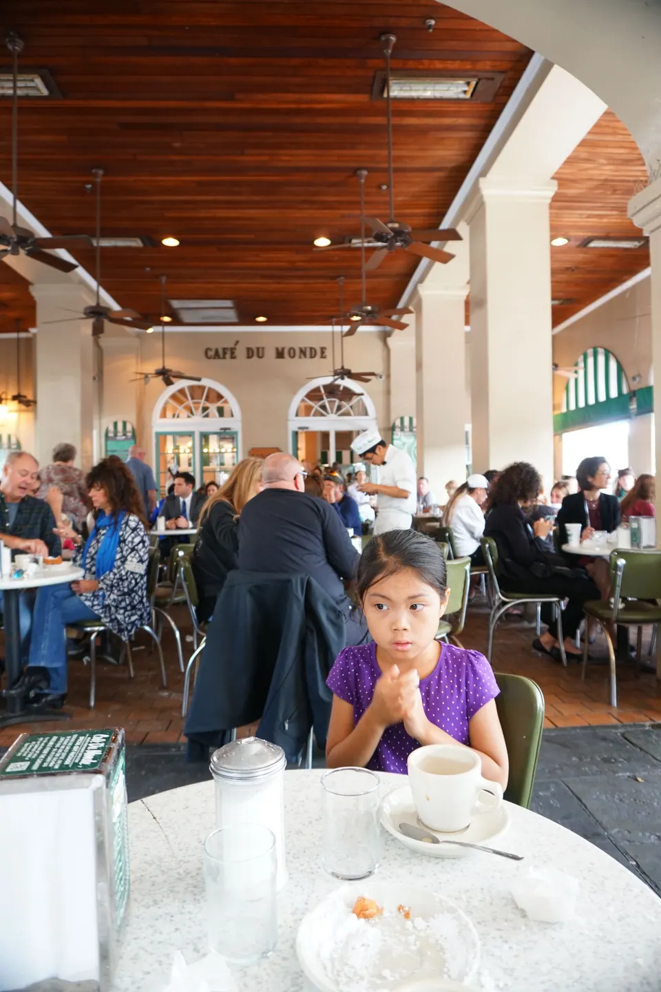 Inside a busy café under ceiling fans, a girl in a purple top sits at a marble table with a coffee cup and a plate dusted with powdered sugar.