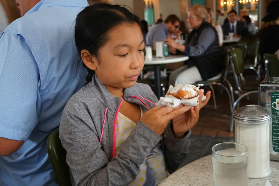 A girl in a grey jacket holds a powdered-sugar beignet in both hands at a café table.
