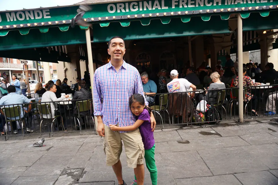 A man in a checked shirt stands in front of the green-and-white Café du Monde awning with a girl in purple hugging his side.