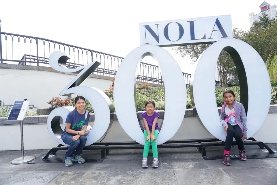 A woman sits on the '3' and two girls sit inside the two zeros of giant white 'NOLA 300' letters in front of a stone wall.