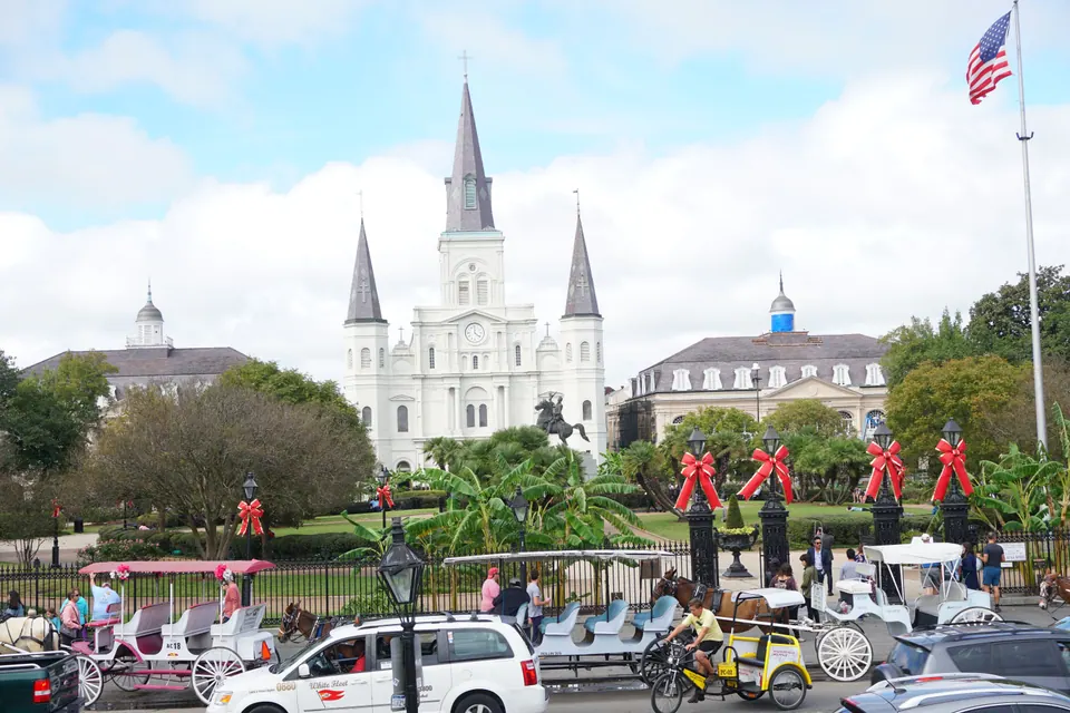 A wide view across a street lined with mule carriages to Jackson Square, with the white three-spired St. Louis Cathedral between two stately buildings and a US flag overhead.