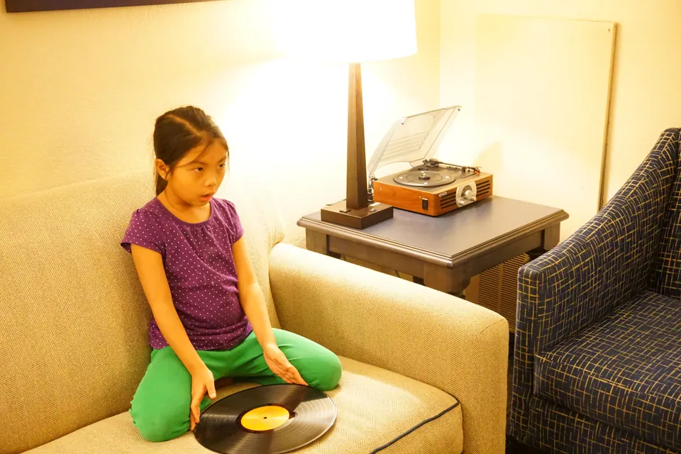 In a warm-lit hotel room, a girl kneels on a sofa with a vinyl record beside her and a small record player on the side table.