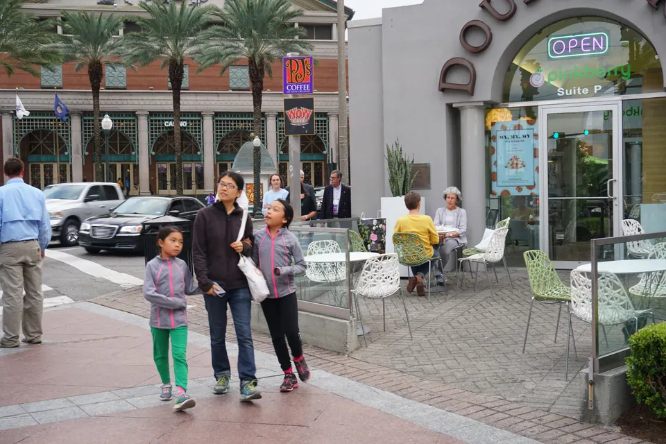 A woman and two girls walk along a sidewalk past a café's outdoor tables, with palms and a large casino building behind.