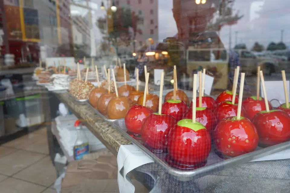 Trays of glossy red candy apples and caramel apples on sticks behind a shop window.