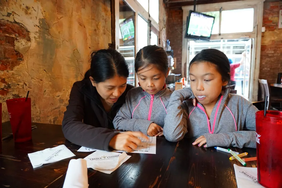 A woman and two girls in matching grey jackets bend over a kids' menu with crayons at a dark wooden restaurant table.