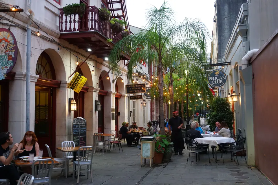 A narrow palm-lined passage with café tables, string lights and a Pelican Club sign at dusk.