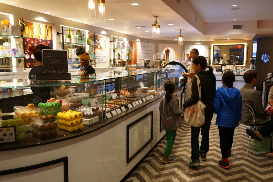 A woman and two girls walk along a long glass counter of macarons and chocolates in a bright sweets shop.