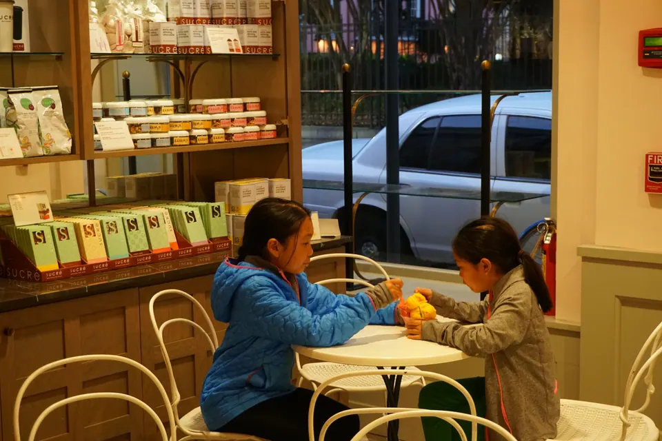 Two girls share a cup of orange gelato at a small white table by a shop window.