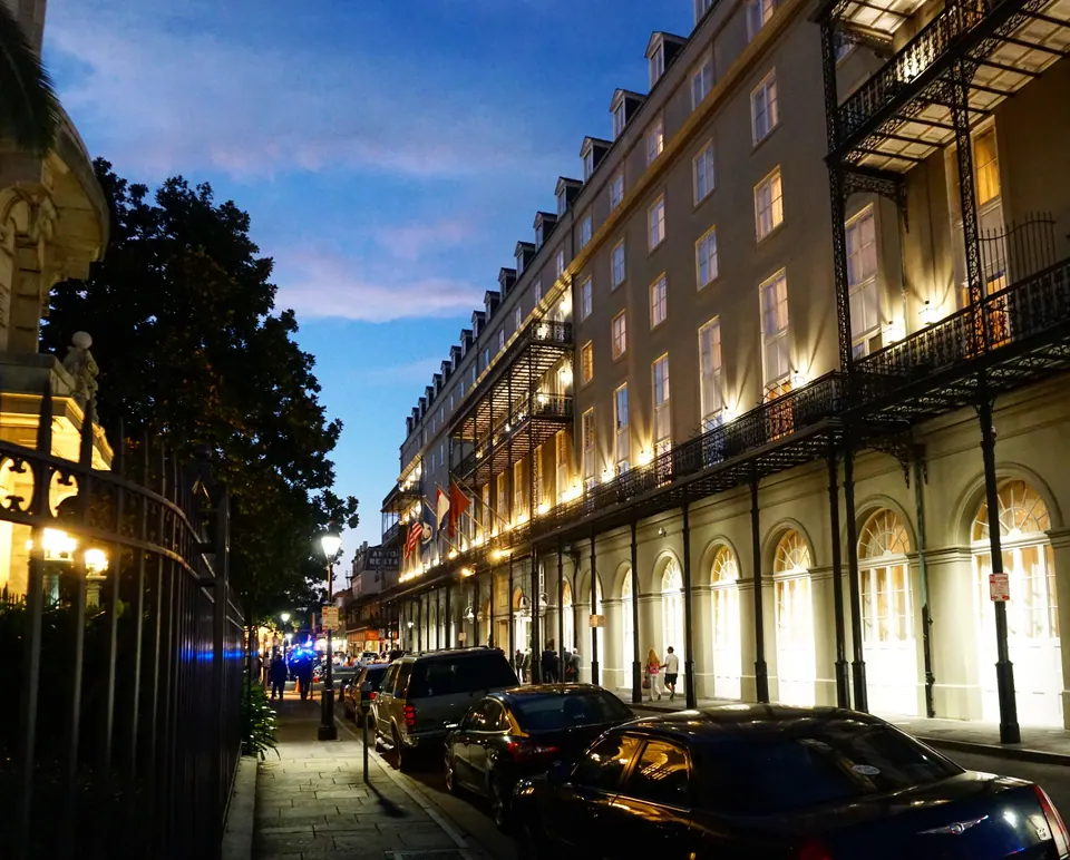 A long lit building with iron galleries glows under a deep blue dusk sky, with cars parked along the street.