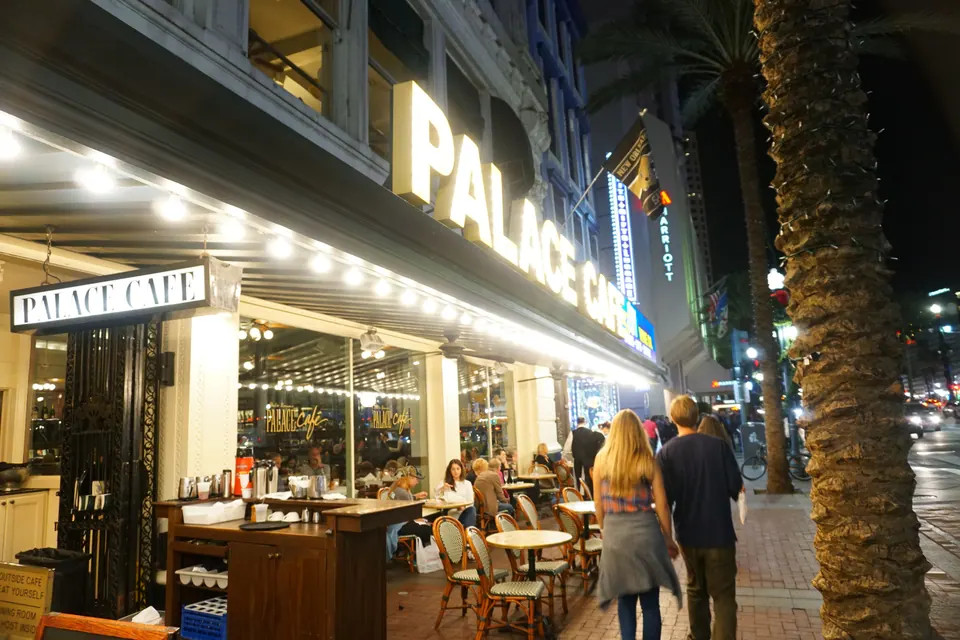 A restaurant's bright marquee reading Palace Café above sidewalk tables on a city street at night.