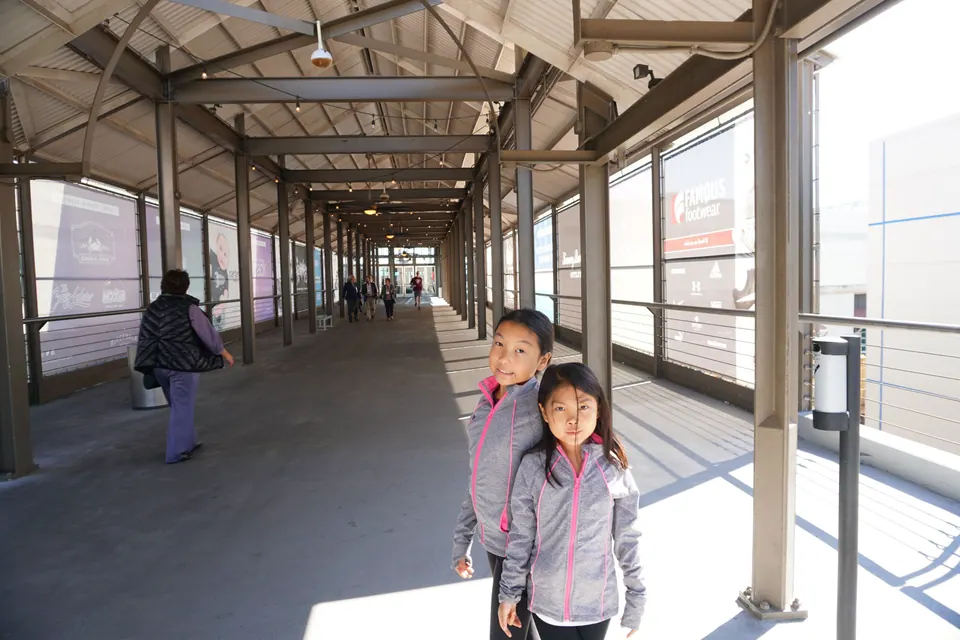 Two girls in matching grey jackets pose in a long, covered walkway with steel columns and glass walls.