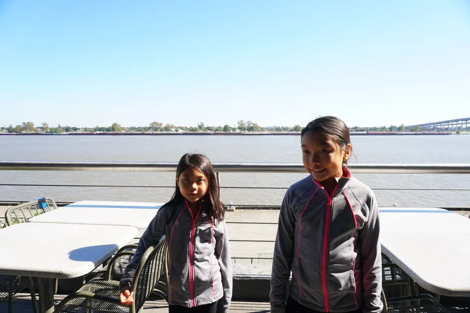 Two girls in grey jackets stand on a sunny terrace with tables, the wide Mississippi River and a distant bridge behind them.