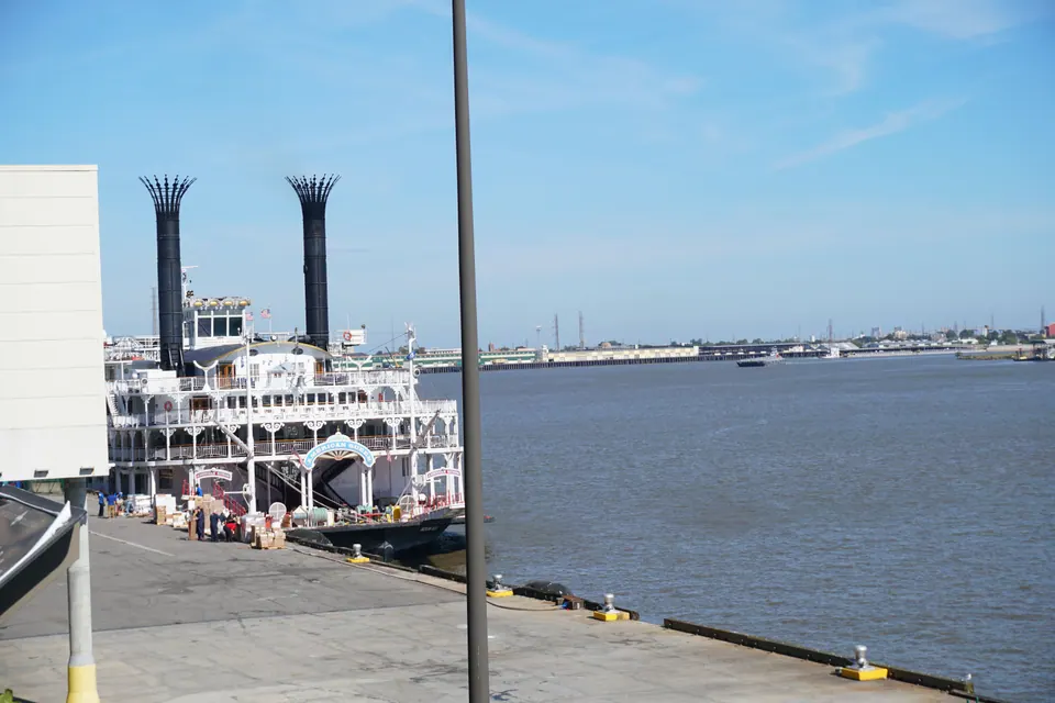 A large white paddle-wheel riverboat with twin black smokestacks docked at a wharf on a wide river under a blue sky.