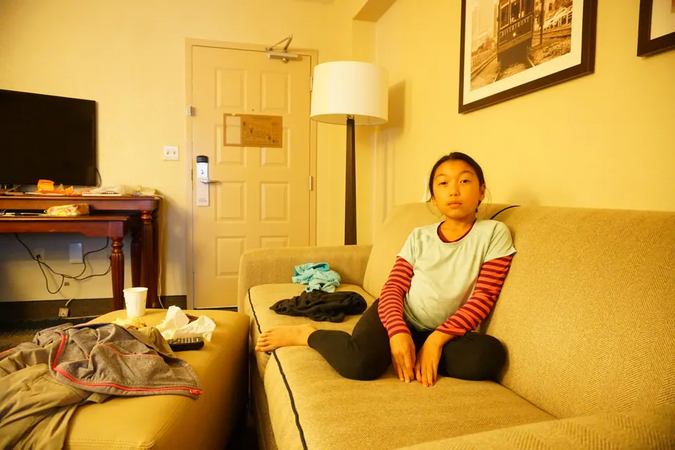 A girl sits cross-legged on the sofa of a hotel suite, a jacket piled on the ottoman beside her.