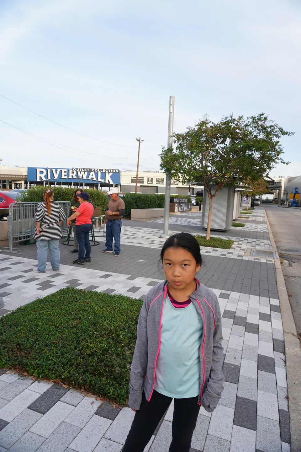 A girl in a grey jacket stands on a paved plaza, with a blue Riverwalk Outlet Collection sign behind her.