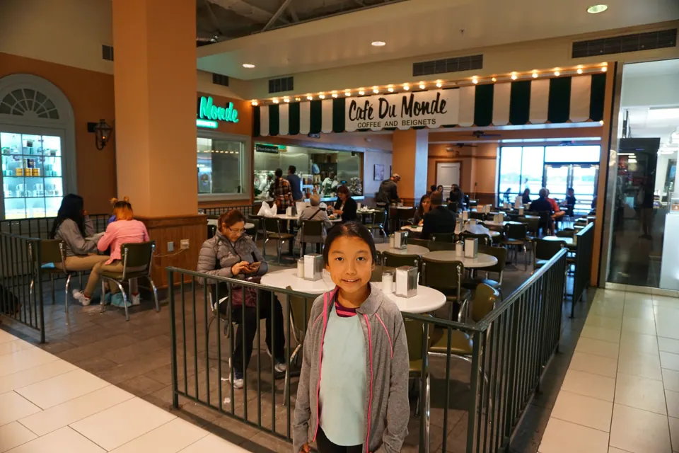 A girl stands at the railing of a Café du Monde counter-service café inside a shopping mall, under its striped awning.