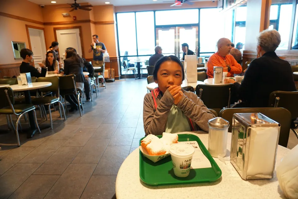 A girl sits behind a green tray of powdered-sugar beignets and a café au lait cup at a round table.