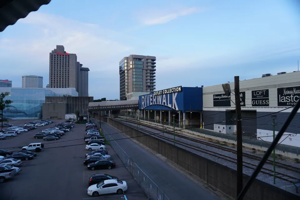 Looking down from a walkway over a parking lot and rail tracks toward the Riverwalk building and downtown towers at dusk.