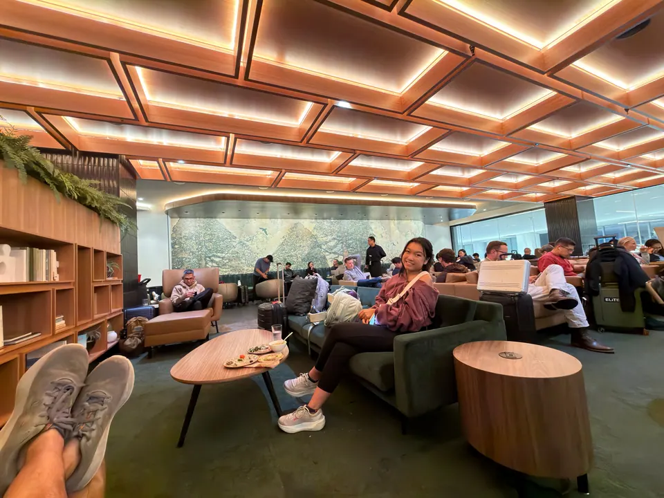 A girl smiles from a green sofa in a busy airport lounge under a glowing wooden coffered ceiling; a pair of sneakers rests on a coffee table in the foreground.