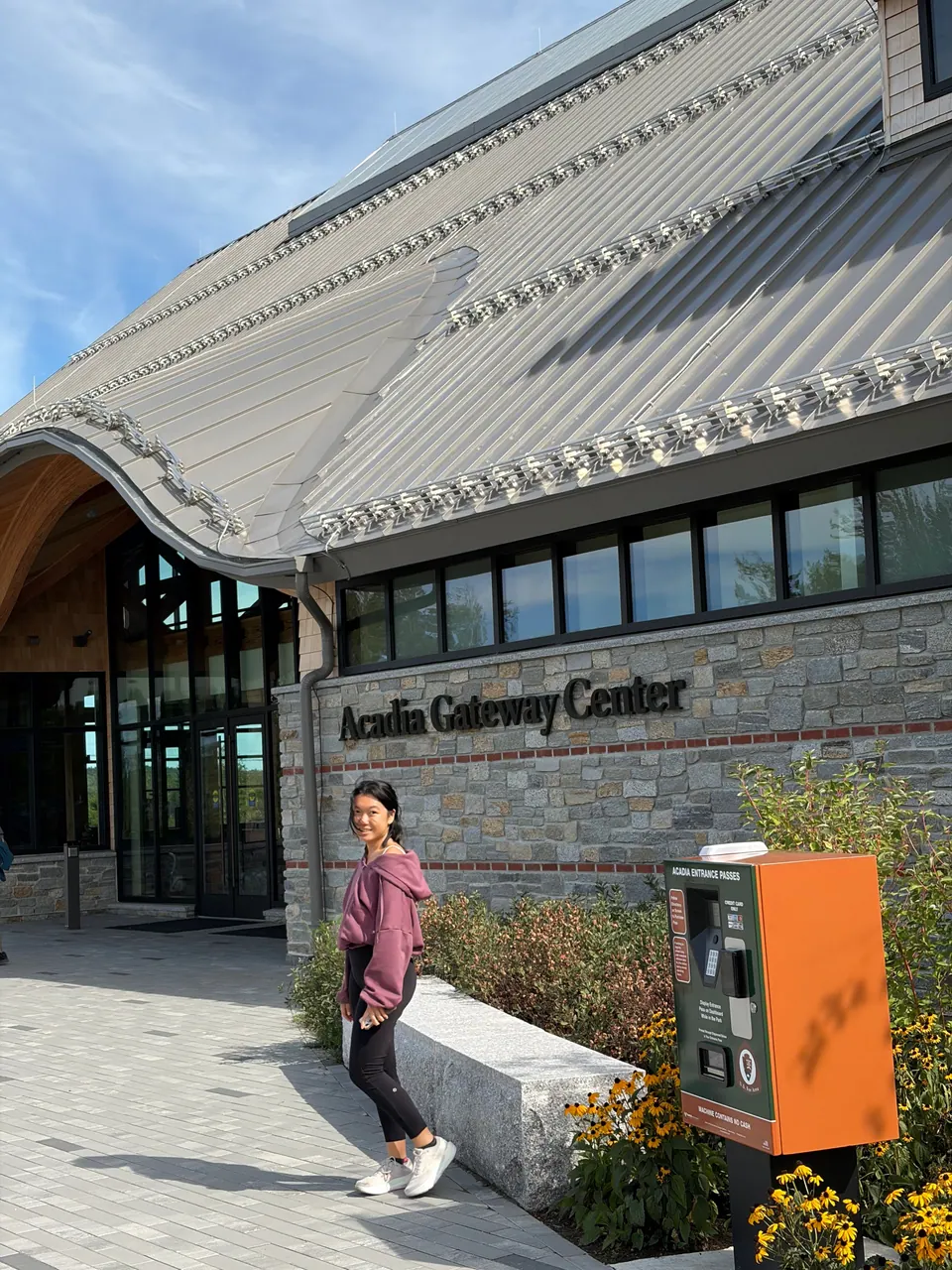 A girl in a mauve sweatshirt stands on a paved walk outside a stone-and-shingle building with a curved metal roof and the sign Acadia Gateway Center; an orange pass kiosk and yellow flowers are in front.