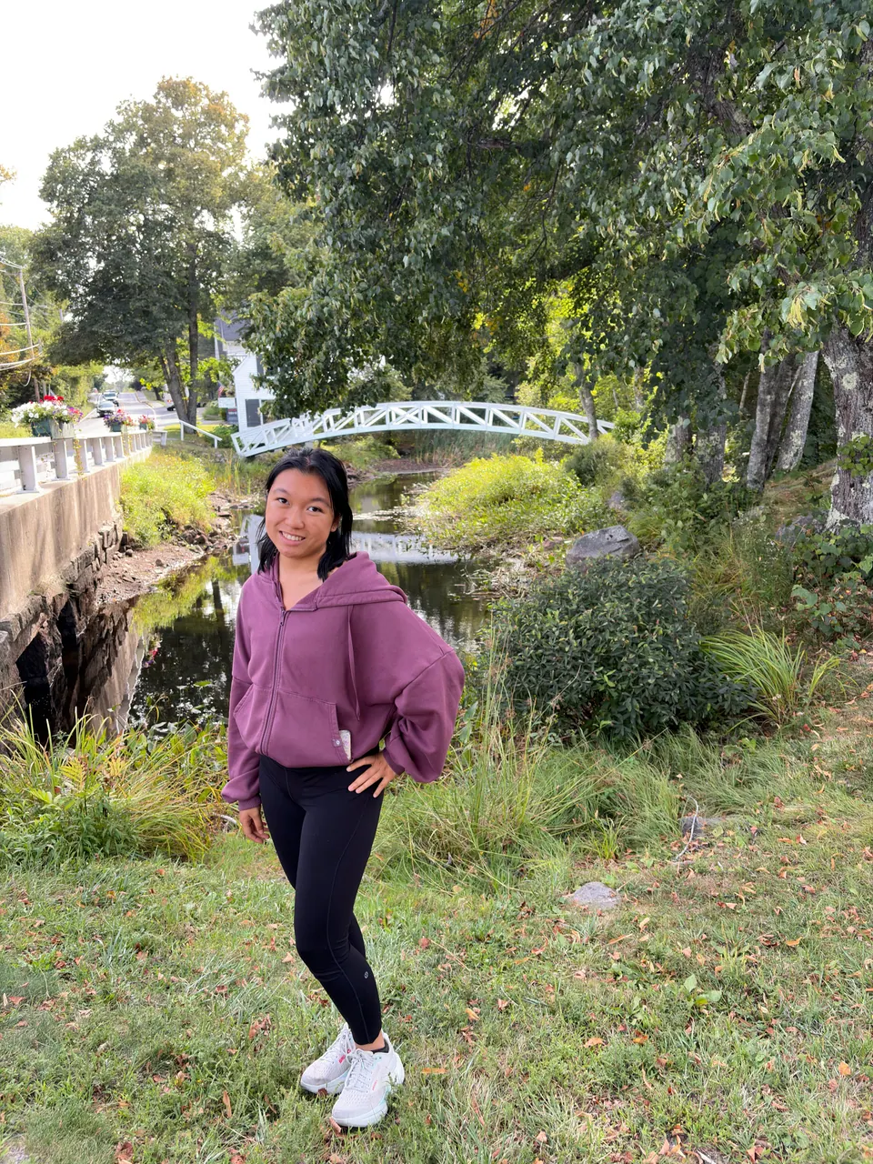 A girl in a mauve zip-up sweatshirt stands on a grassy bank by a small stream, with a white arched wooden footbridge among trees behind her.