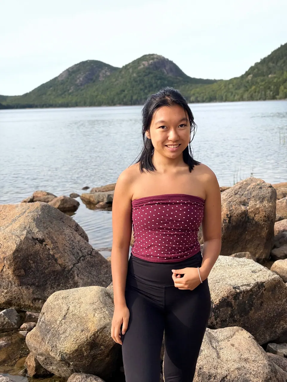 A girl in a dotted red top stands among granite boulders at the edge of a lake, with two rounded wooded hills across the water.
