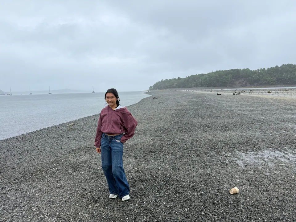 A girl in a maroon sweatshirt and jeans stands on a wide wet gravel bar under a grey sky, with calm water on the left, a wooded island behind and sailboats in the distance.