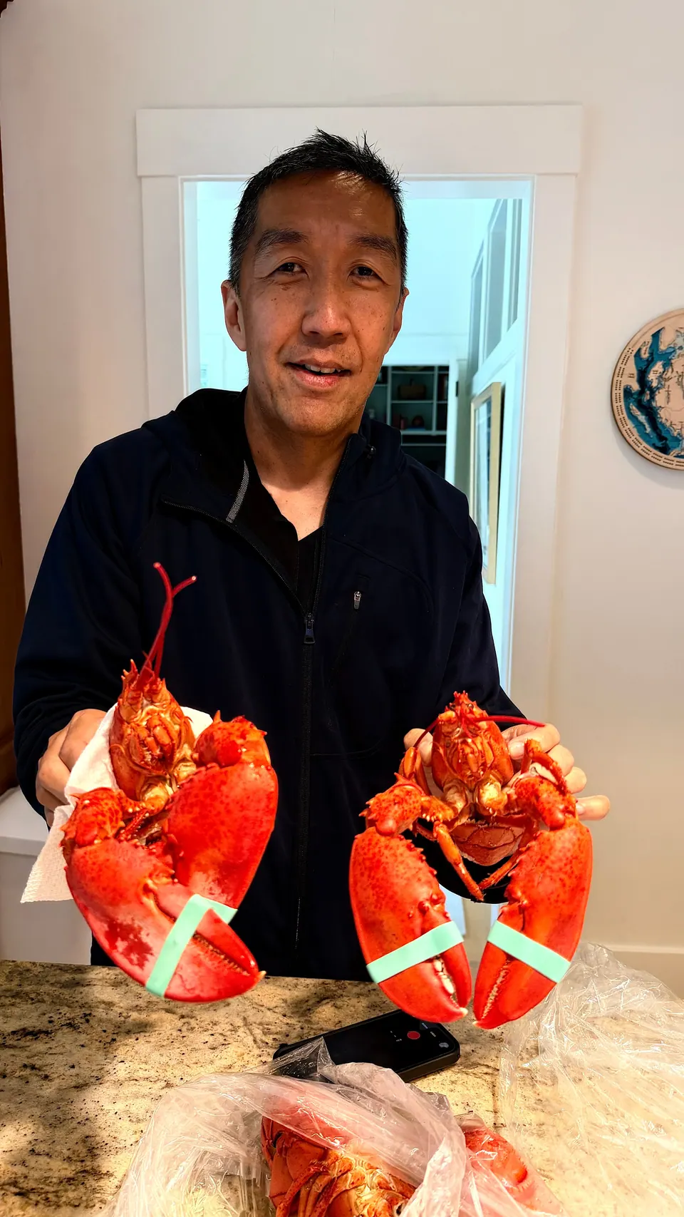 A smiling man in a dark jacket holds up two bright red cooked lobsters with banded claws over a granite kitchen counter.
