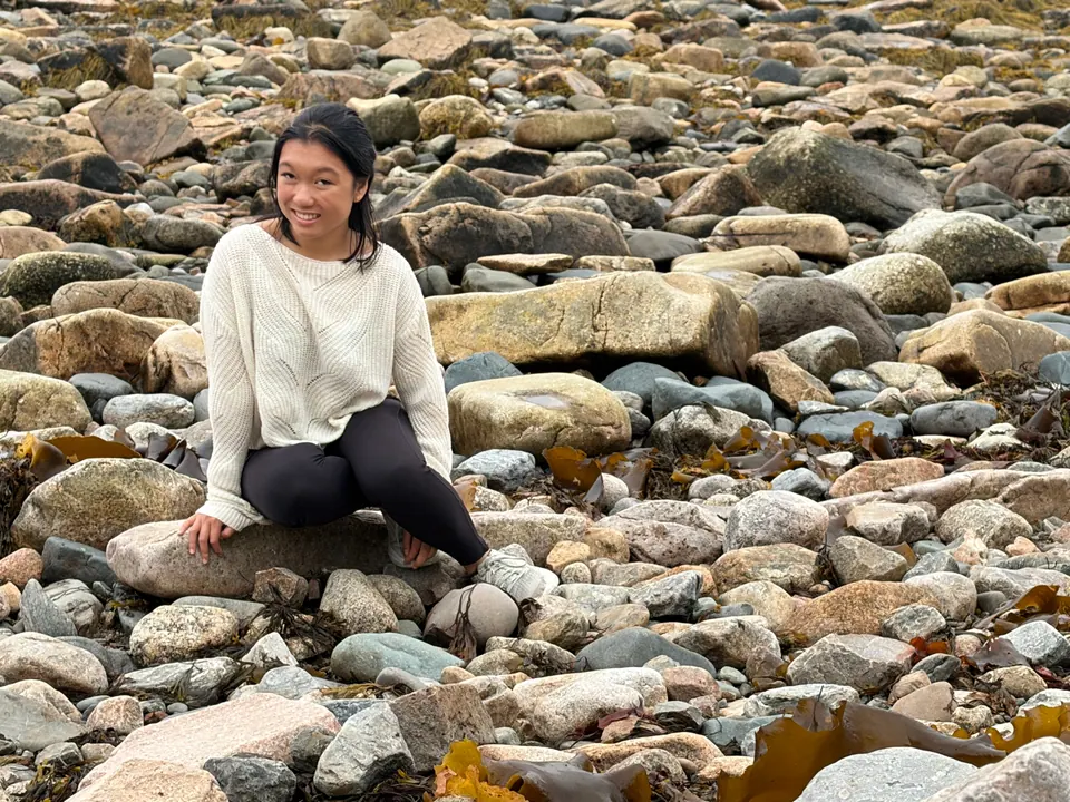 A girl in a cream sweater sits smiling on a beach of rounded grey and tan stones and boulders, with seaweed among them.